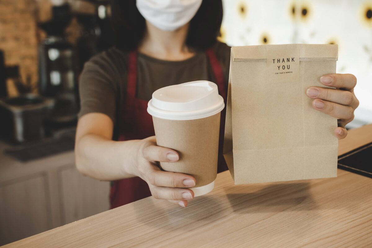 Masked restaurant worker handing a takeout coffee cup and bag, symbolizing restaurant adaptation to COVID restrictions.